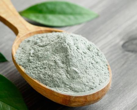 Close-up of a cosmetic dry gray clay in a wooden spoon on a black wooden table. Decorated with green leaves.